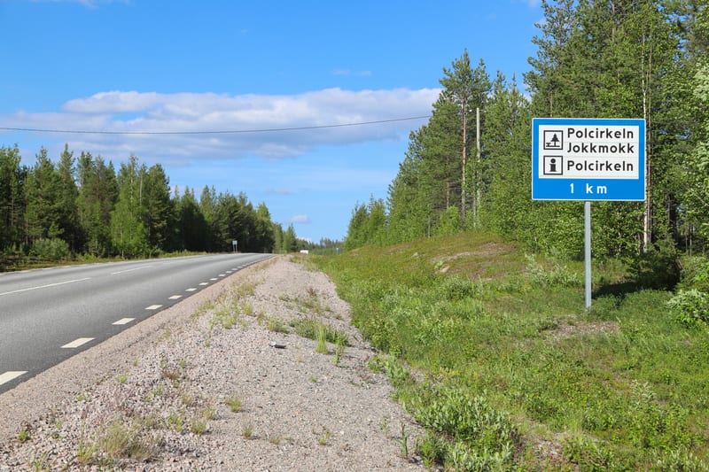 Bilden visar en asfalterad väg med en delvis grusbelagd vägren som sträcker sig i bildens vänstra del. Längs med vägen, på höger sida, finns en blå skylt med vit text som indikerar "Polcirkeln", "Jokkmokk" och "1 km". I bakgrunden och längs sidorna a