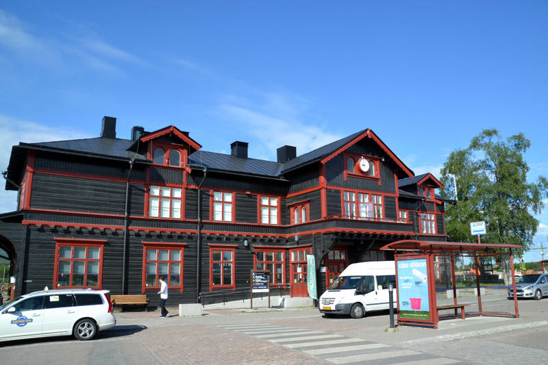 Bilden visar en exteriör av en byggnad med mörka fasader och röda detaljer vid fönster och taklister. Byggnaden har flera fönster och takkupor, samt en klocka på fasaden. Framför byggnaden finns ett vitt fordon, möjligen en taxi, och en person som gå
