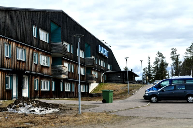 Bilden visar fasaden på en flervåningsbyggnad klädd med mörkt trä, delvis med vertikala och horisontella brädor. Byggnaden har flera vita fönster och balkonger med trädetaljer. På den mörka delen av fasaden syns en ljus skylt med texten "DUNDRET". Fr