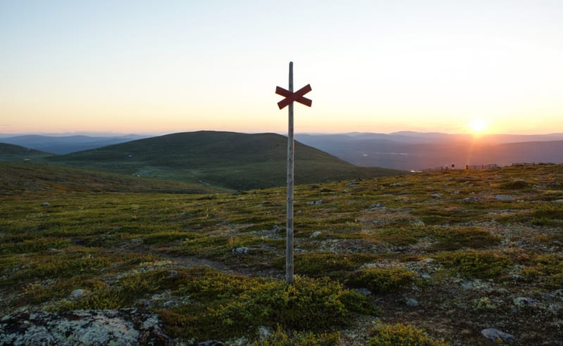 En bild som visar en markerad vandringsled i ett fjällandskap under solnedgång eller soluppgång. I mitten av bilden står en trästolpe med ett rött kryss som markerar leden. Marken är täckt av grön vegetation, mossa och stenar. Bakgrunden består av ku