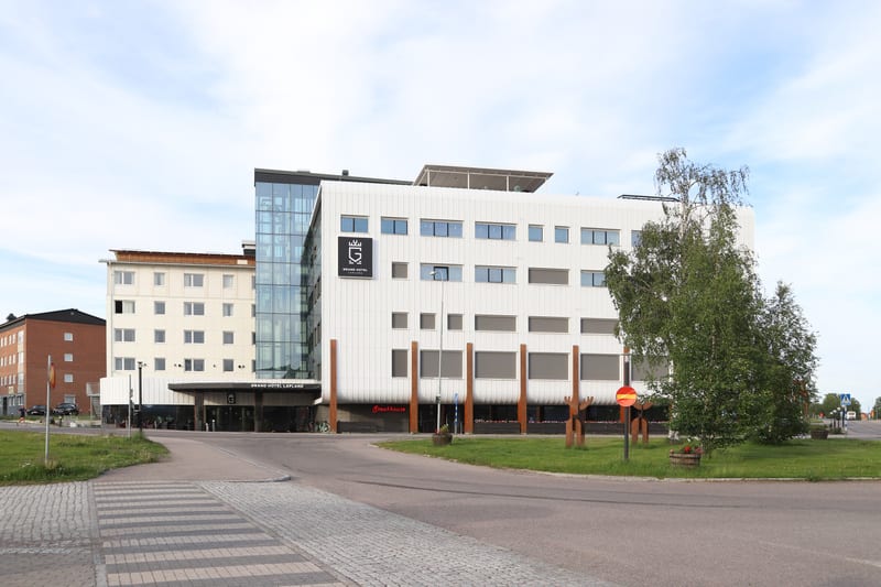 En bild utomhus som visar Grand Hotel Lapland och dess omgivning under dagtid. Hotellet består av flera sammankopplade byggnader, varav en är ljusbeige med många fönster och en annan är vit med varierande fönsterstorlekar och en stor glasfasad. Entré