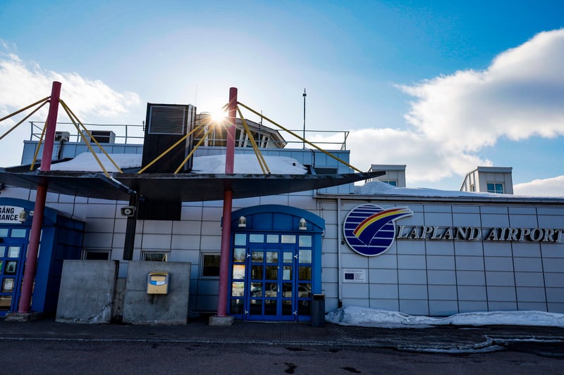 Bilden visar utsidan av en byggnad med skylten "LAPLAND AIRPORT" på fasaden. Byggnaden har en modern design med grå paneler, blå dörrar och detaljer i rött och gult. Ett tak täckt med snö täcker delar av entréområdet, och mer snö syns längs byggnaden