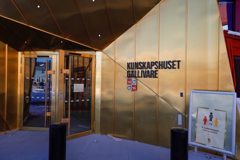 Bilden visar entréområdet till "KUNSKAPSHUSET GÄLLIVARE". Fasaden är täckt med blanka gyllene eller kopparfärgade paneler som är vinklade. Två glasdörrar med guldlist är synliga, varav den högra är något öppen. I reflektionen från dörrarna syns en lj
