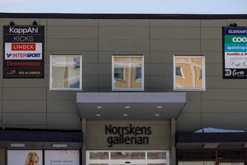 Bilden visar fasaden på en byggnad, sannolikt ett köpcentrum eller en galleria, under dagtid. Fasaden är grå med flera rutor och skyltar för olika butiker och tjänster. Framträdande skyltar inkluderar 'KappAhl', 'KICKS', 'Lindex', 'Intersport', 'Dres