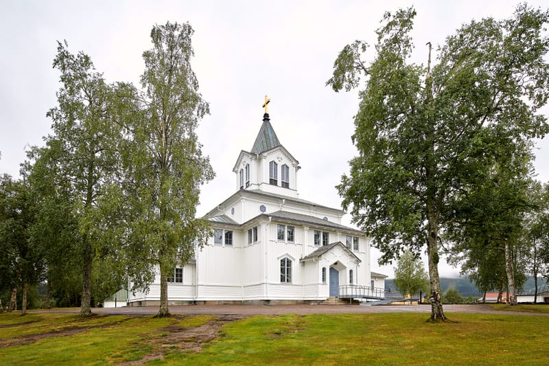 Bilden visar en vit kyrkobyggnad med en spira, toppad med ett gyllene kors, under en gråmulen himmel. Byggnaden är omgiven av gräsmatta och flera lövträd med gröna blad, troligen björkar, varav några står framför kyrkan. På vänster sida av kyrkan led