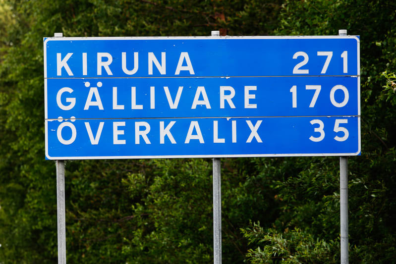 En blå vägskylt med vit text som visar avstånd till tre orter. Överst står 'KIRUNA 271', i mitten 'GÄLLIVARE 170' och nederst 'ÖVERKALIX 35'. Skylten är monterad på två metallstolpar och bakom den syns grön växtlighet, troligen träd eller buskar. Tex