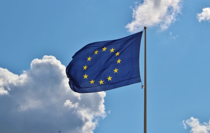 Bilden visar Europarådets flagga, ofta kallad EU-flaggan, som vajar mot en blå himmel med vita moln i bakgrunden. Flaggan är blå med tolv gyllene stjärnor arrangerade i en cirkel. Den är fäst vid en flaggstång som syns på höger sida av bilden. Solen