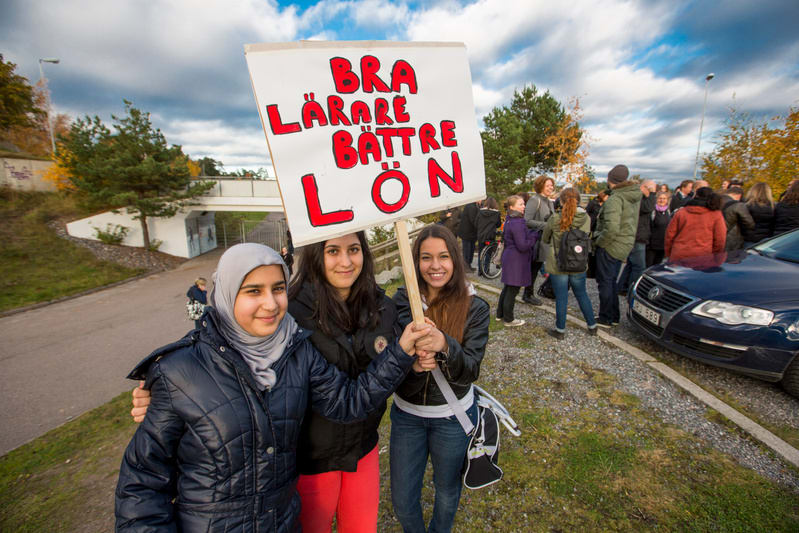 Bilden visar tre unga kvinnor i förgrunden som håller upp en skylt med texten "BRA LÄRARE BÄTTRE LÖN". En av kvinnorna bär en grå hijab. Kvinnorna ler mot kameran. Bakom dem syns en folksamling med flera personer, en mörkblå bil är delvis synlig till