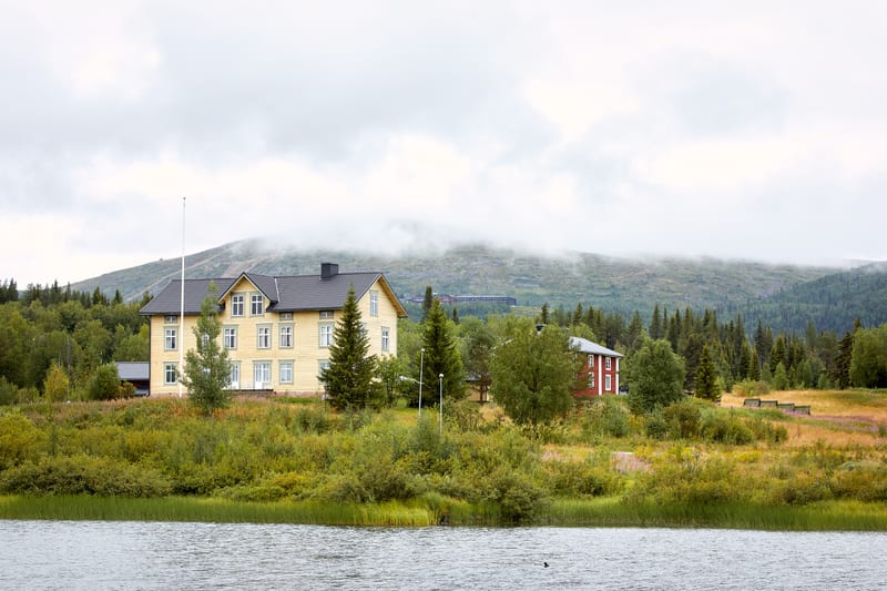 Bilden visar ett landskap med en sjö eller större vattensamling i förgrunden. På strandkanten i mitten av bilden finns ett stort, ljust gult hus med vit trim och ett mörkt tak. Bredvid det gula huset står två barrträd. Längre bort till höger syns ett