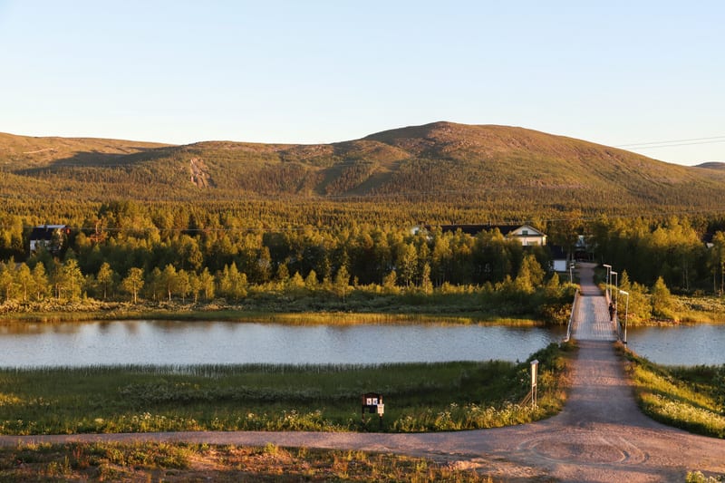 En bild visar ett landskap med en sjö eller större vattendrag i förgrunden, som reflekterar den ljusa himlen. Över vattnet sträcker sig en träbro med metallräcken, som leder till en bebyggelse. Runt vattnet och mot bron finns grönskande vegetation, i