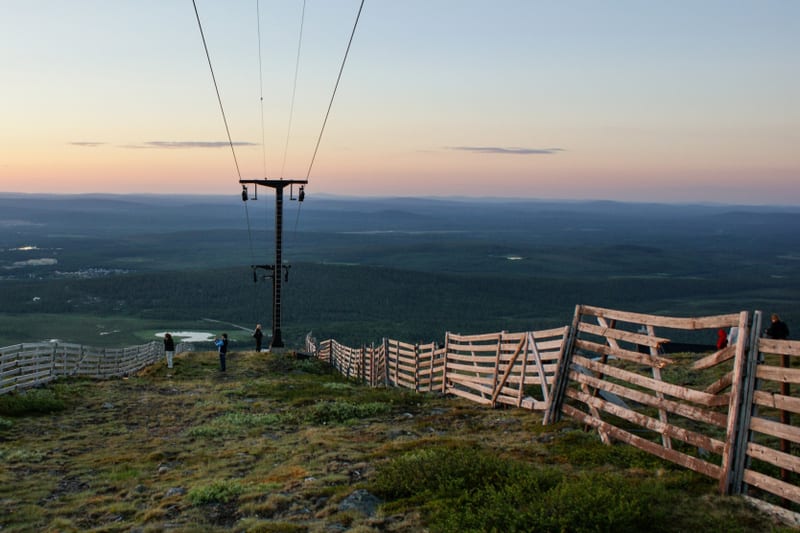 Bilden visar ett landskap taget från en höjd, troligen ett fjäll, vid skymning eller gryning. Himmelen är upplyst i toner av blått, rosa och orange. I förgrunden syns gräs och små buskar, samt en del stenar. En trästaket sträcker sig diagonalt över b