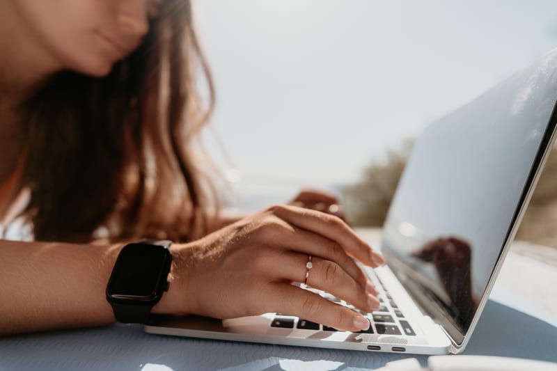 En bild tagen utomhus under dagtid, sedd från en låg vinkel. I förgrunden syns en person som använder en laptop. Personens ansikte är oskärpt och det mesta av fokus ligger på händerna och laptopen. En hand med en ring på ringfingret och en svart smartklocka på handleden vilar på tangentbordet. Personens långa, mörkbruna hår syns på höger sida av bilden. Ljuset kommer uppifrån höger, vilket skapar starka kontraster och ljusfläckar. Bakgrunden är ljus och oskärpt, vilket indikerar att det är soligt väder.