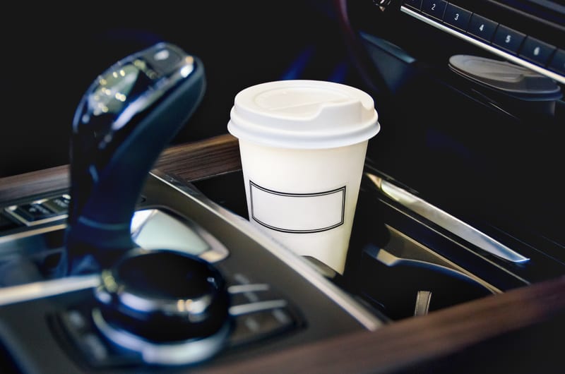 Bilden visar interiören av en bil, med fokus på mittkonsolen. I mitten står en vit pappersmugg med ett vitt lock och en tom rektangulär etikett på framsidan. Till vänster om muggen syns delar av en automatisk växellådsspak och en kontrollratt. Till höger om muggen syns en del av bilens instrumentbräda med knappar märkta '1', '2', '3', '4', '5', med mera, samt en yta med reflekterande metalliska detaljer. Trädetaljer ramar in delar av mittkonsolen. Bakgrunden är mörk och oskarp.