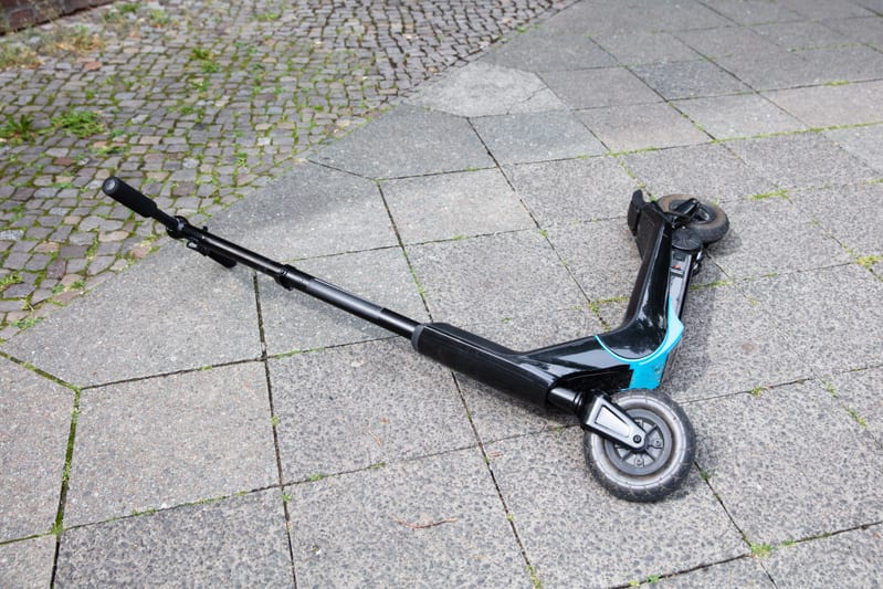 En svart sparkcykel med turkosa detaljer ligger på marken på en gångväg. Sparkcykeln är vänd upp och ner med styret riktat åt vänster och hjulen synliga. Gångvägen består av grå betongplattor med inslag av gräs eller mossa i fogarna. Till vänster i bilden syns en yta med kullerstenar och lite mer gräs. Ljuset indikerar en utomhusmiljö.
