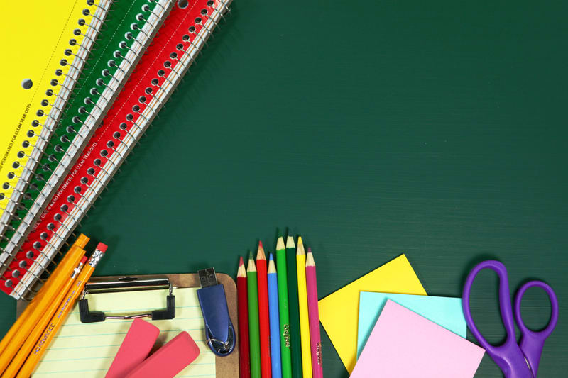 Bilden visar en samling skolmaterial arrangerade mot en mörkgrön bakgrund, som påminner om en skoltavla. I det övre vänstra hörnet ligger tre spiralblock i färgerna gult, grönt och rött. I bildens nedre vänstra del syns flera gula blyertspennor, en k
