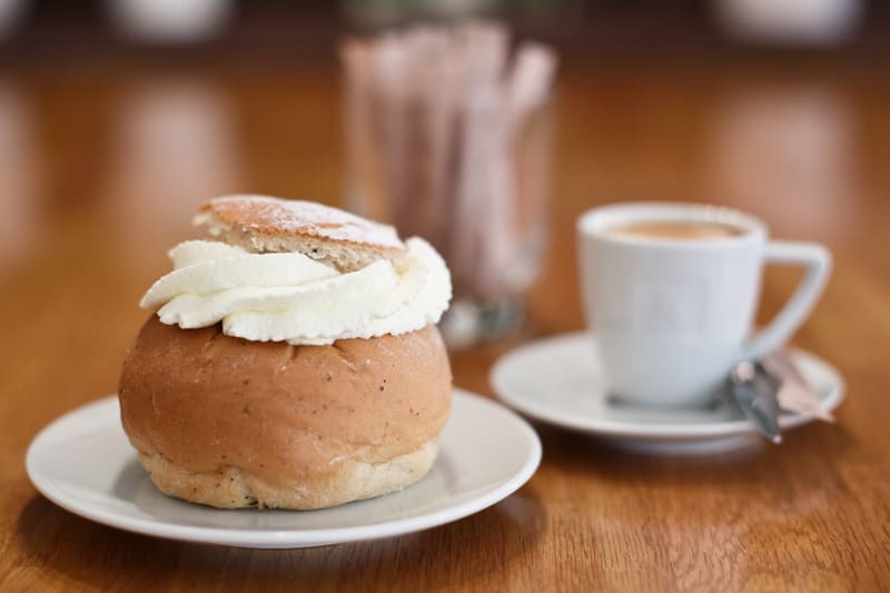 En semla med grädde och locket på toppen, serverad på ett vitt fat, står i förgrunden till vänster i bilden. Semlan är pudrad med florsocker. Till höger om semlan står en vit kaffekopp på ett matchande vitt tefat, innehållande en mörk dryck, troligen