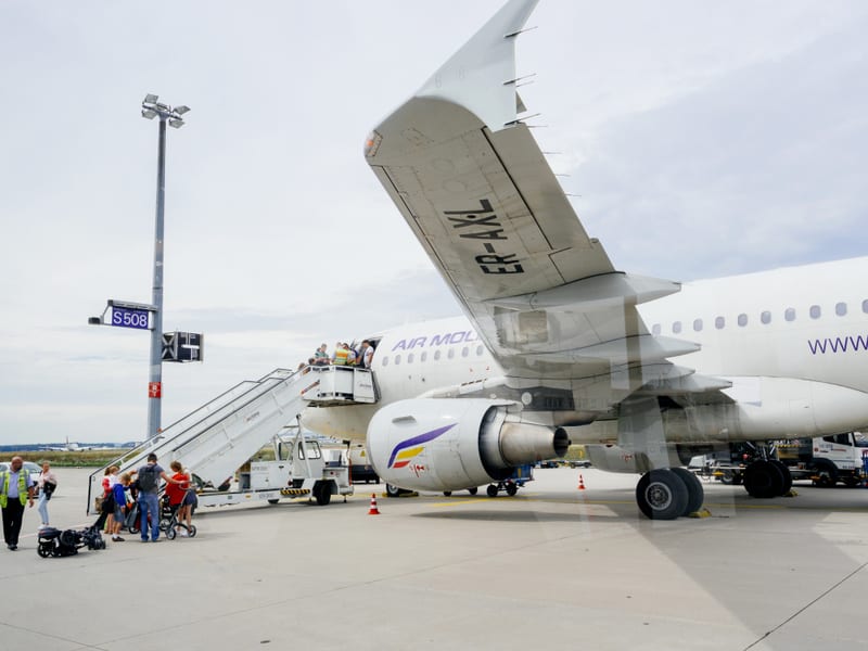 Bilden visar ett passagerarflygplan av typen Airbus A320, delvis synligt, med flera personer som går ombord via en passagerartrappa. Flygplanet är vitt med lila text där "AIR MOLDOVA" och "WWW.AIRMOLDOVA.MD" kan utläsas. Registreringsnumret "ER-AXL"