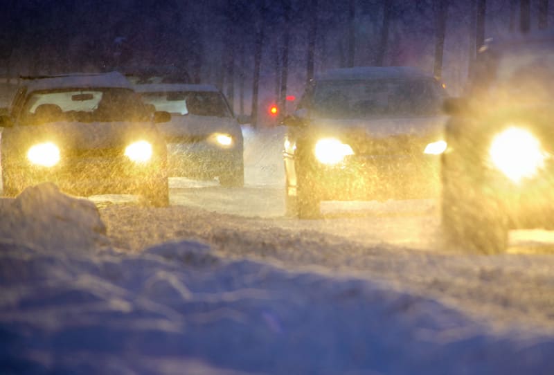 En bild tagen i mörker av flera bilar som kör under kraftigt snöfall. Vägen är täckt med snö och snödrev syns i bilarnas strålkastarljus. Fyra bilar är synliga; tre som kör mot betraktaren och en som kör bortåt. Bilarna som kör mot betraktaren har tä