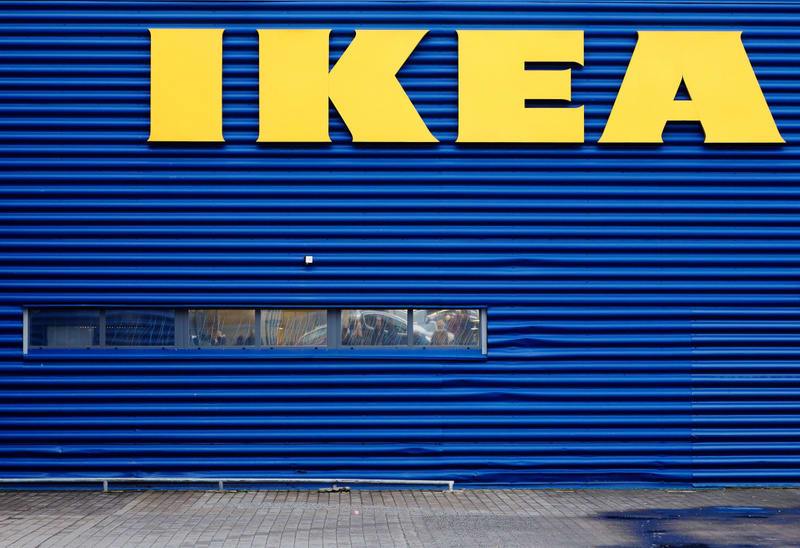 Bilden visar fasaden på en byggnad, som är klädd i horisontella, korrugerade blå plåtar. Högst upp i bilden, placerat centralt, syns den gula logotypen 'IKEA'. Nedanför logotypen och till vänster om mitten av fasaden finns en horisontell fönsterrad med flera rutor. Genom fönstren kan man se otydliga reflektioner och skuggor som skulle kunna vara människor eller föremål inomhus. Längst ner i bilden syns en markyta av gråa markstenar, som delvis är våta och reflekterar ljus.