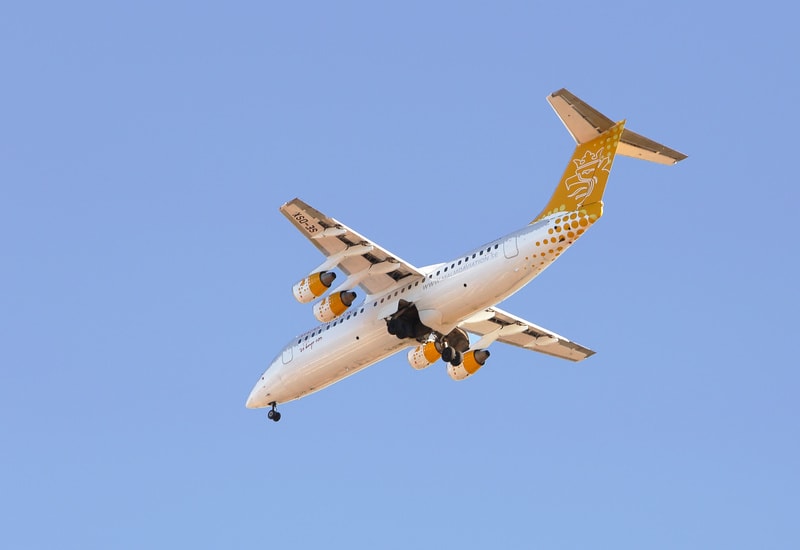 Bilden visar ett passagerarflygplan, fotograferat underifrån mot en klarblå himmel. Flygplanet har en vit flygkropp med gula detaljer på vingen och stjärtfenan. Stjärtfenan har en gul krönliknande logotyp och en design med gula prickar. Undersidan av vingen har texten "SE-OSX" och på flygkroppen syns "WWW.HALMDIAVIATION.SE". Landing gear (landningsställ) ser ut att vara utfällt. Fyra motorer är synliga under vingarna, med gula inslag på motorhöljen.