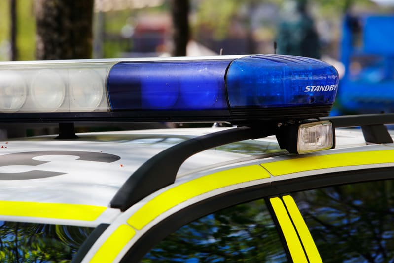 Bilden visar ovansidan av en polisbil i detalj, med fokus på taket och varningsljusrampen. Varningsljusrampen är delvis vit och delvis blå, med texten "STANDBY" synlig på den blå delen. En extra vit strålkastare är monterad bredvid den blå delen av rampen. På taket av fordonet är en svart takräcke synlig. Bilens kaross är vit med gula ränder längs taklinjen och på dörrkarmen. Reflektioner av träd och himmel syns svagt i bilens fönster. Bakgrunden är oskarp med antydningar av grönska och andra föremål.