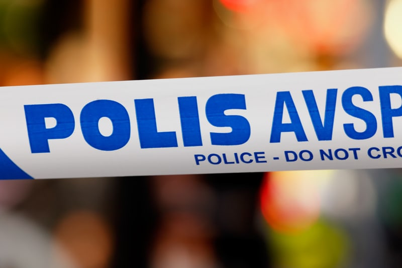 Bilden visar ett nära perspektiv på en vit avspärrningstejp med blå text. Texten "POLIS AVSP" är tydligt synlig i stor stil, och därunder står den mindre texten "POLICE - DO NOT CRO". Tejpens diagonala ränder i blått och vitt syns också. Bakgrunden är oskarp med suddiga färgade ljuspunkter, vilket antyder att avspärrningstejpen är framträdande i förgrunden medan bakgrunden är utanför fokus. Ljuspunkterna i bakgrunden är i nyanser av rött, orange, gult och möjligtvis grönt, vilket kan indikera lampor eller belysning i en urban miljö.