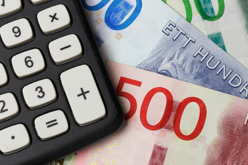 En närbild av en svart räknare med vita knappar som är placerad diagonalt över tre svenska sedlar. De synliga delarna av räknaren är tangentbordet med numeriska knappar (3, 6, 9) och matematiska operationsknappar (multiplikation, subtraktion, addition, lika med och punkt). Sedlarna som ligger under räknaren är delvis synliga och visar valörer som 500 och 100, samt texten 'ETT HUNDRA'. Färgerna på sedlarna är varierande, med inslag av rött, blått och grönt. Ljuset kommer från övre högra hörnet och skapar en skarp skugga från räknaren över sedlarna.