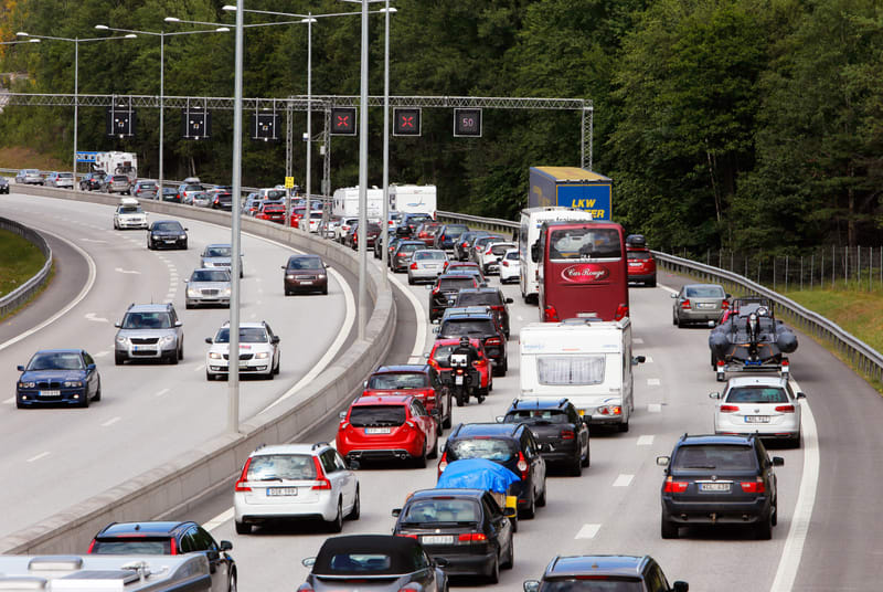 Bilden visar en motorväg med intensiv trafik. Flera körfält är fyllda med fordon som rör sig långsamt eller står stilla, vilket indikerar en bilkö. I förgrunden och mellanregistret syns personbilar, husvagnar, en röd buss och en lastbil. På en del bi
