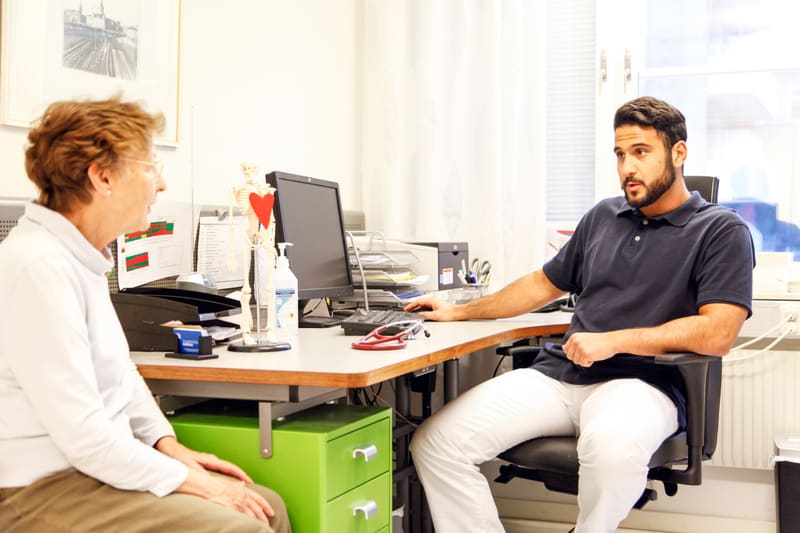 Bilden föreställer en man och en kvinna som sitter i ett rum, troligtvis ett kontor eller mottagningsrum. Kvinnan, som är på vänster sida, sitter vänd mot mannen och har brunt hår och glasögon. Hon bär en vit långärmad tröja. Mannen, som sitter på hö