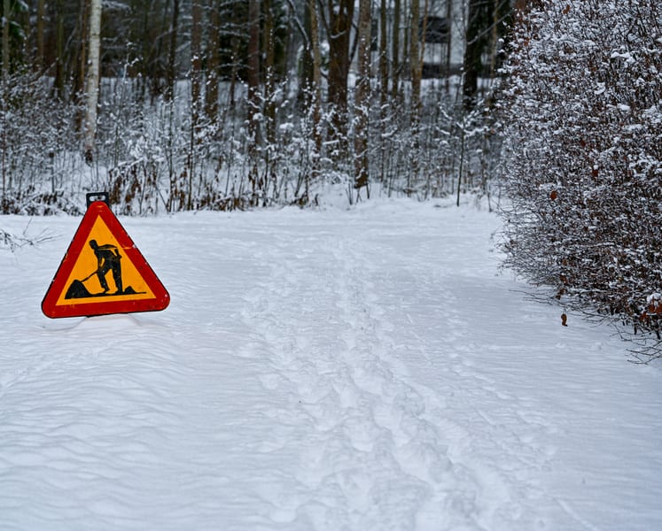 En bild tagen utomhus visar en snötäckt mark med ett varningsskylt i förgrunden till vänster. Skylten är triangulär med röd ram, gul bakgrund och en svart siluett av en person som skottar eller gräver med en spade. Spår, troligen från fötter, syns i snön som leder bortåt i bilden. Till höger i förgrunden finns en buske med grenar och några enstaka torra löv, delvis täckt av snö. I bakgrunden syns träd, även de täckta med snö, och delar av vad som verkar vara byggnader skymtar längre bort genom träden.