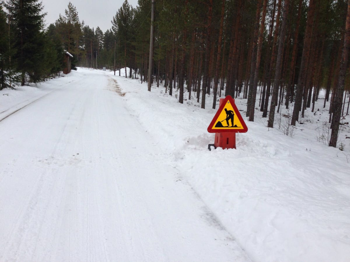 Bilden visar en snötäckt grusväg som sträcker sig in i en skog. Längs vägkanten, till höger i bild, står en varningsskylt för vägarbete. Skylten är röd och gul, triangulär, och visar en svart silhuett av en person som skottar. Skogen består primärt a