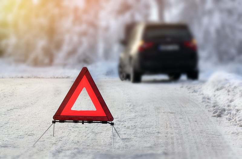 En röd varningstriangel står uppställd på en snötäckt väg i förgrunden, med fokus på triangeln. I bakgrunden syns en mörk personbil, vars bakdel är otydlig på grund av oskärpa. Vägen är täckt av snö och det syns spår av fordon. Till höger om vägen li