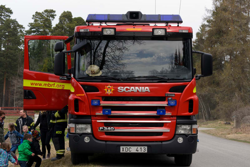 Bilden visar fronten av en röd brandbil av märket Scania. På brandbilens dörr, som står öppen, syns texten "www.mbrf.org" på en gul remsa. En gul brandhjälm med numret "32" ligger på bilens instrumentbräda, tydligt synlig genom vindrutan. Bilen har b
