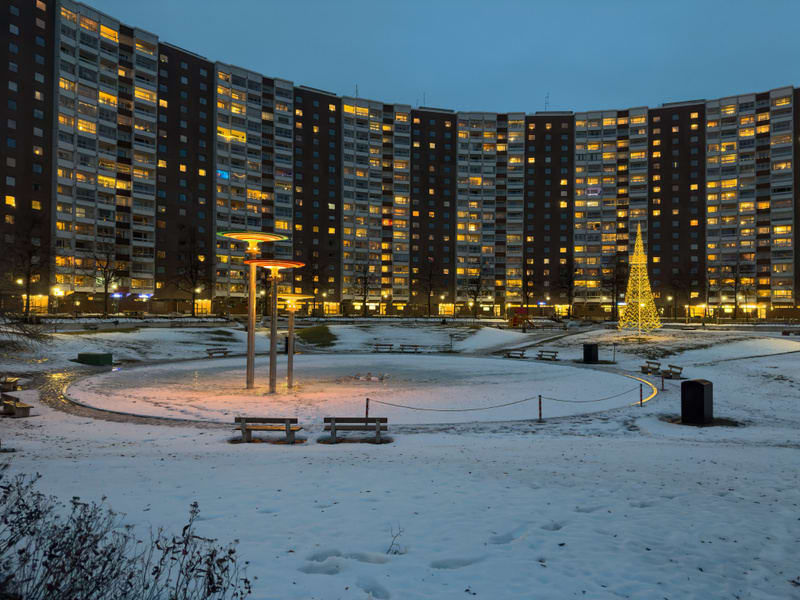 Bilden visar ett snötäckt grönområde framför en stor, kurvad byggnad med många upplysta fönster. Grönområdet har snö på marken och en halvcirkelformad asfalterad gångväg i mitten. Flera parkbänkar är placerade runt gångvägen. Det finns också tre pela