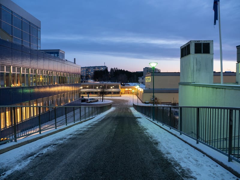 Bilden visar en utomhusmiljö under skymning eller gryning, med ett snötäckt landskap. I förgrunden syns en asfalterad väg eller gångväg, delvis täckt av snö och is, som lutar nedåt. Längs vägen finns metallräcken på båda sidor. Till vänster i bilden