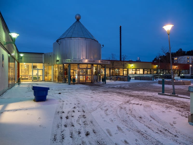 Bilden visar ett snötäckt torg eller en öppen yta framför en byggnad med flera ingångar. Marken är täckt av snö med spår av däck och fotsteg. Till vänster finns en blå soptunna. Byggnaden har stora glaspartier och ljusa väggar. En central del av bygg