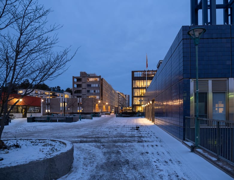 Bilden visar ett stadstorg eller en öppen plats täckt med snö under kvällstid eller gryning, med blått skymningsljus i himlen. Till vänster i förgrunden syns en bar trädgren. På vänster sida av bilden finns flera byggnader, inklusive en med en röd yt