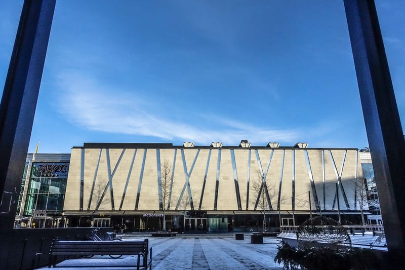 En bild tagen utomhus under dagtid som visar en byggnad med en fasad av ljusa paneler och diagonala silverfärgade detaljer. Framför byggnaden finns ett snötäckt torg med rutor av ljusa och mörkare plattor. Längst ner till vänster i bilden syns en trä