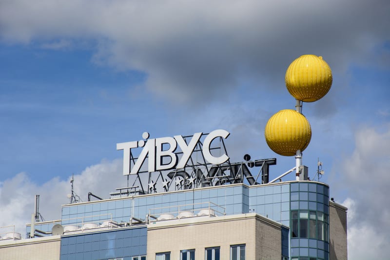 Bilden visar toppen av en byggnad med en stor skylt med texten "TÄVUC". Skylten är placerad på taket av en flervåningsbyggnad som har delar klädda i blåaktiga glaspaneler och delar i ljusare tegel eller puts. Ovanför skylten, till höger, finns två st