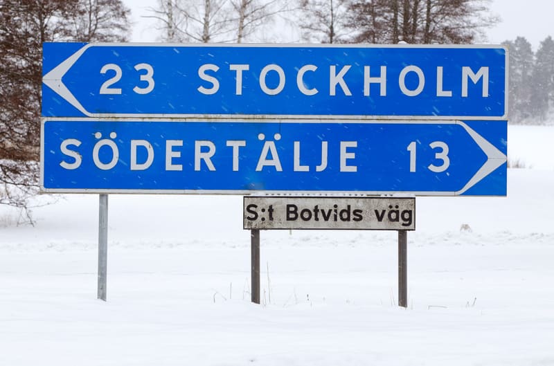 Bilden visar tre skyltar i ett snötäckt landskap med träd i bakgrunden. Den översta skylten är blå med vit text som anger "23 STOCKHOLM" och har en vit pil som pekar åt vänster. Den mellersta skylten är också blå med vit text och anger "SÖDERTÄLJE 13