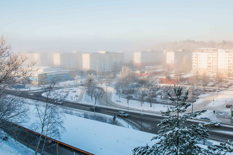 En översiktsbild av ett vinterlandskap som visar en stadsmiljö täckt av snö. I förgrunden syns snötäckta träd och buskar, med en sluttning som leder ner mot en väg. På vägen kör flera bilar. I mitten av bilden finns en byggnad med delvis blå fasad oc