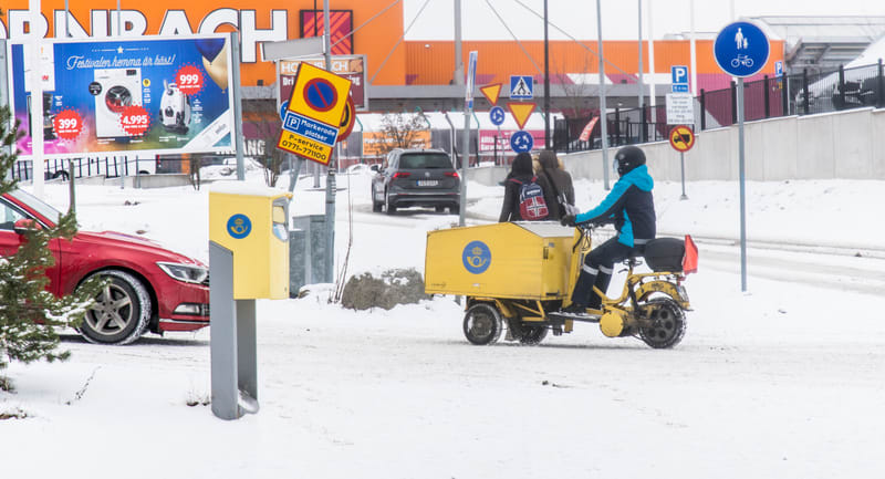 Bilden visar ett landskap täckt av snö, vilket indikerar vinterförhållanden. I förgrunden till vänster syns en röd personbil delvis täckt av snö. Bredvid bilen står en gul brevlåda. Centralt i bilden är en postmoped av äldre modell, också gul, med en