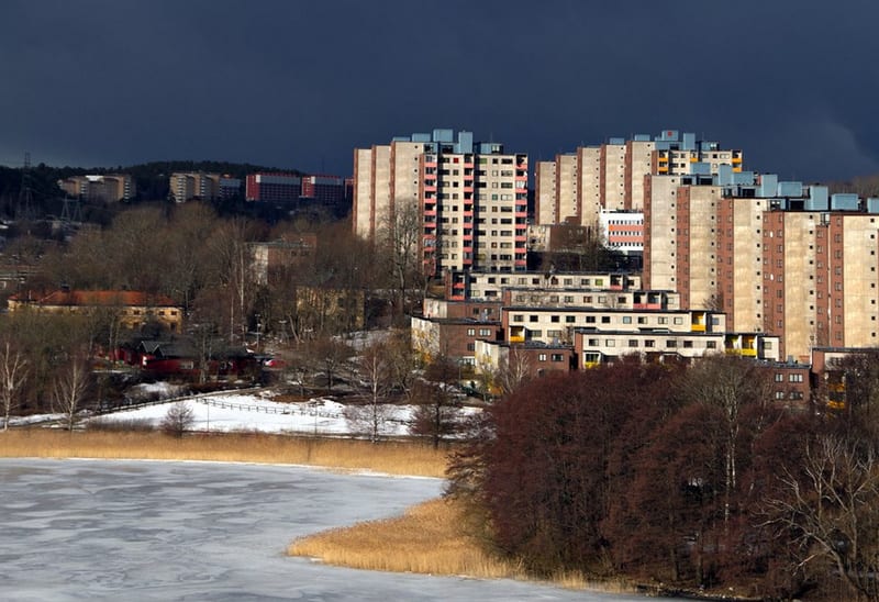 Bilden visar ett landskap med flera höghus och lägre byggnader i förgrunden och på avstånd. Hälften av bilden upptas av en isbelagd sjö med vass vid stranden, med snötäckta områden närmare husen. Träd, de flesta lövfällande, står kala eller med höstf
