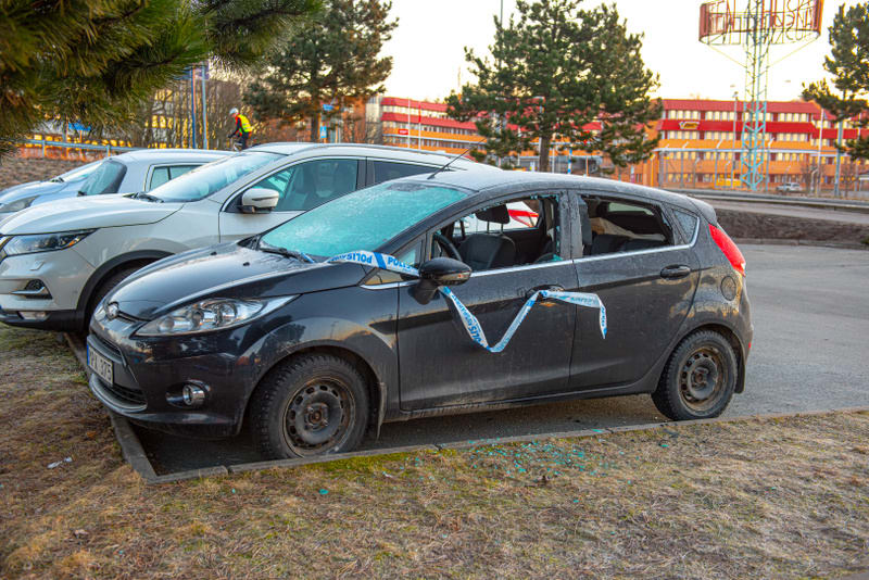 Bilden visar en svart fyrsitsig personbil som står parkerad på en gräsbeklädd yta bredvid en trottoarkant. Bilens vänstra sidoglas är krossade och öppna, och den högra sidorutan är också krossad. Bilens framruta är täckt av frost eller is, och en vit