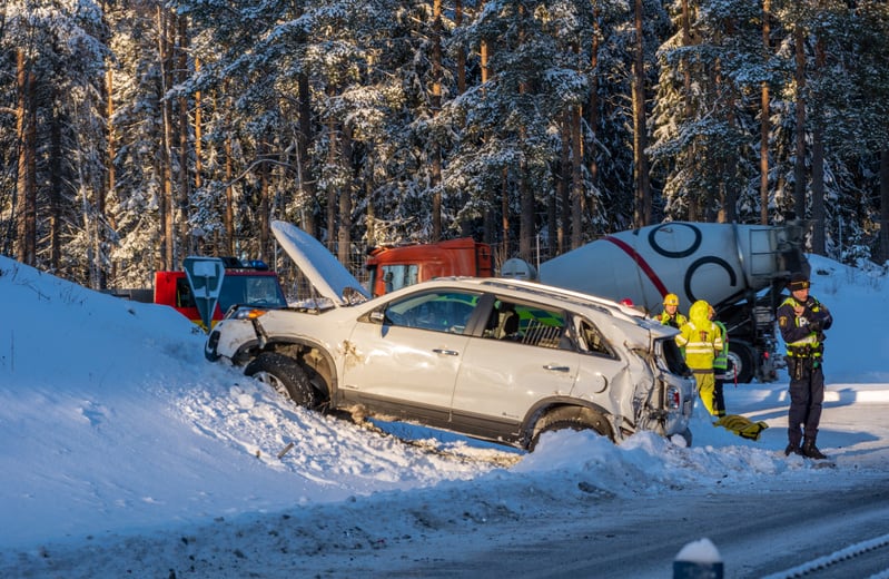 Bilden visar en trafikolycksplats under vintertid vid sidan av en väg, omgiven av snötäckta träd. En vit personbil står delvis nere i ett snötäckt dike med fronten skadad och motorhuven öppen. Bakre delen av bilen är också skadad, och bilen är omgive
