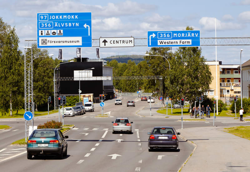 En bild tagen dagtid utomhus som visar en vägkorsning med trafik och flera vägvisningsskyltar. Två stora blå skyltar hänger över vägen med information om vägnummer (97, 356) och destinationer som Jokkmokk, Älvsbyn, Morjärv, samt Forsvarsmuseum och We