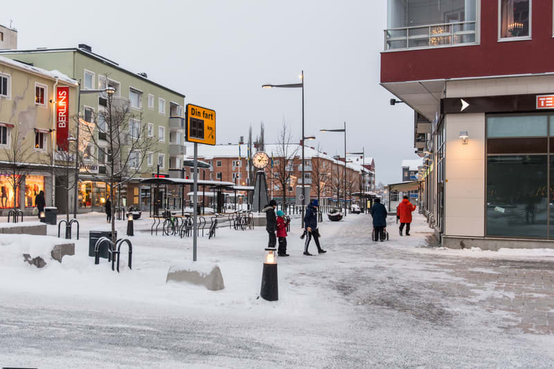 En vinterbild tagen utomhus från en gata eller torg i en stad. Mark och hustak är täckta av snö. Till vänster syns flera flerfamiljshus, varav ett har en röd skylt med texten "BERLINS". På höger sida finns en modern byggnad med stora fönster och en r