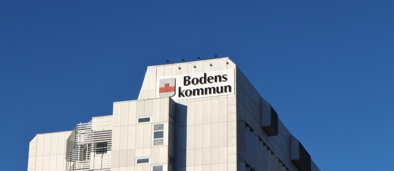 Bilden visar toppen av en modern byggnad under en klarblå himmel. På byggnadens tak, mot himlen, står en stor skylt med texten 'Bodens kommun' och Breds kommunvapen, som är en röd borg på vit bakgrund. Byggnaden är vit eller ljusgrå med flera fönster