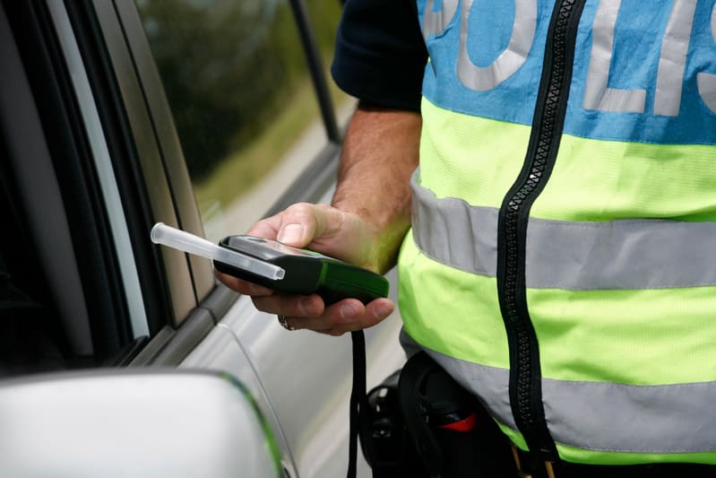 Bilden visar närbild på en person klädd i en reflexväst med texten "POLIS" på överdelen. Personen håller en alkomätare i höger hand, med munstycket riktat mot ett fordons sidoruta. Fordonet är silverfärgat och delar av fordonets dörrhandtag, backspeg