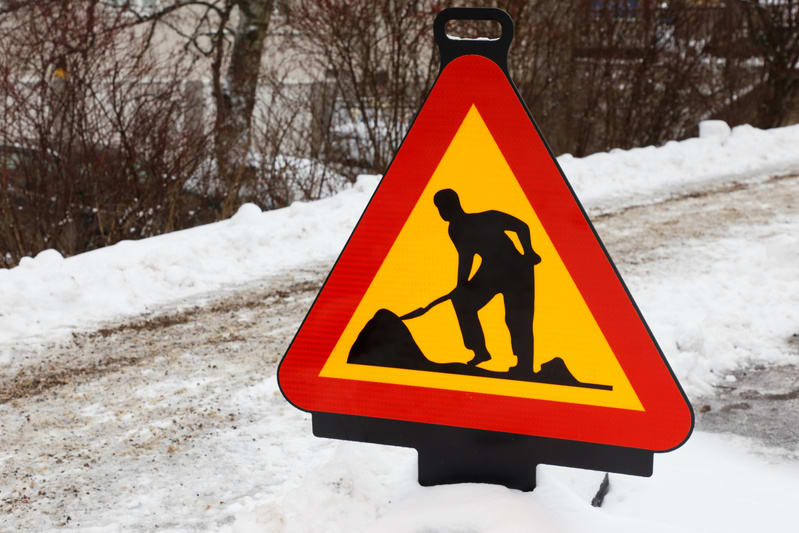 En gul varningsskylt med röd ram står placerad i snöig terräng. Skylten visar en svart silhuett av en person som gräver med en spade. Snön på marken är vit med smutsiga fläckar av gråbrun mark, och den sträcker sig upp längs en slänt i bakgrunden. Et